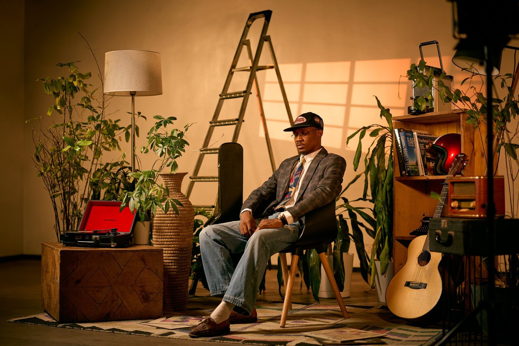 Sunkanmi Stephens seated in a studio filled with plants, a ladder and an acoustic guitar